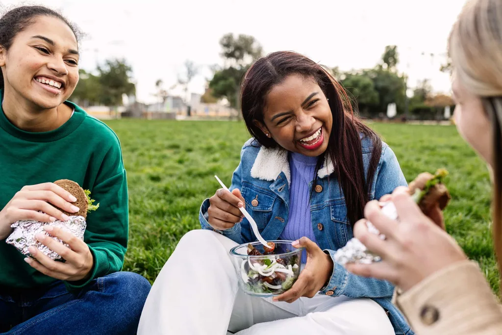 Three young women laughing together while eating lunch on a grassy lawn outdoors, one holding a sandwich and another eating a salad from a glass bowl.