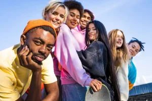 A diverse group of seven young friends smiling and posing together outdoors against a blue sky, one holding a skateboard