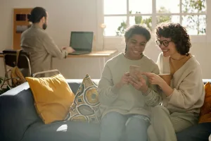 Two people sitting together on a couch, smiling as they look at a smartphone, with a third person working on a laptop in the background.